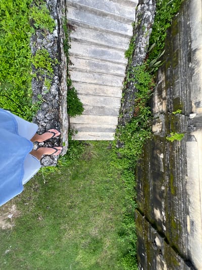 A high-angle forensic shot captures a view looking down into a stone stairwell in Pecatu, Indonesia. A person's bare feet, adorned with black flip-flops and partially obscured by light blue shorts, are positioned at the upper left edge of a rough-hewn stone wall. Below and extending diagonally from the upper center, a series of worn concrete steps descends between two vertical structures. The immediate left structure is a dark stone wall heavily covered with vibrant green foliage. The opposing right structure is a darker, possibly ancient or decorative carved stone wall, exhibiting weathering, discoloration, and mossy patches. A section of green grass is visible at the bottom of the stairwell, directly beneath the observer and to the right of the steps.