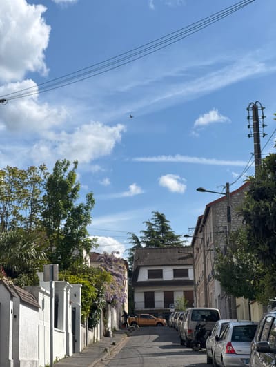 A street in Champigny-sur-Marne, France, is lined with white buildings and greenery. Cars are parked along the side of the road. Overhead wires stretch across the sky with scattered clouds and contrails. A lamppost is visible.