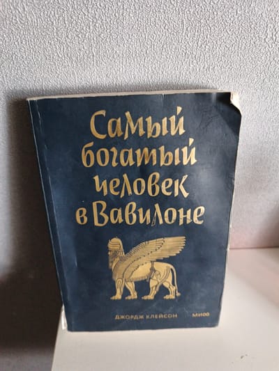 The image shows the cover of a book with a dark blue background and gold lettering. The title reads "Самый богатый человек в Вавилоне," which translates to "The Richest Man in Babylon." Below the title is an illustration of a winged lion. At the bottom of the cover, the text "ДЖОРДЖ КЛЕЙСОН" (George S. Clason) and "МИФ" (Myth) are visible. The book is placed on a white surface, possibly a shelf or table, against a textured, light-colored wall. The overall scene is well-lit and appears to be an indoor setting. There is no information in the image that indicates the location is in Shymkent, Kazakhstan.