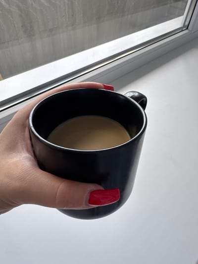 A close-up shot shows a hand with bright red nail polish holding a black mug filled with a light brown liquid, likely coffee or tea. The mug is tilted, revealing the surface of the beverage. The background is divided between a light-colored wall with diagonal shadows and a window frame with light streaming through, illuminating a floor with a grey, tiled pattern. The overall lighting suggests it might be daytime, with sunlight entering from the window. The scene evokes a sense of a quiet, personal moment, possibly during a break or while enjoying a drink by a window.