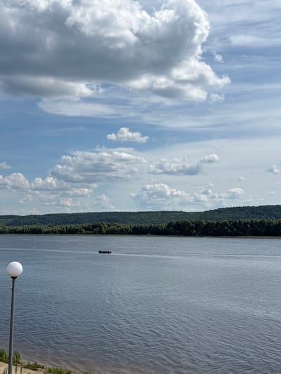 This image depicts a wide river with a distant, tree-covered hillside on the opposite bank. The water is calm with a slight ripple. In the middle ground, a small, dark boat with a single occupant is moving from left to right, leaving a wake. In the foreground on the left, a tall, slender lamp post with a spherical white light fixture stands next to a sandy embankment with sparse green vegetation. The sky is a mix of blue sky and white and gray clouds, some of which are quite large and dramatic. The scene appears to be a natural landscape, likely a riverbank or park area, within or near the city of Zvenigovo, Russia, given the context provided.