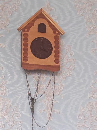 A wooden cuckoo clock with a dark brown face and Roman numerals hangs on a light blue wall with an ornate, light-colored pattern. The clock has a house-shaped top with a dark brown square opening above the clock face. The clock face has Roman numerals indicating the time is approximately 10:55. Two metal chains hang from the bottom of the clock, disappearing out of frame. The image appears to be taken indoors. There is no information to indicate the location in the city of Homyel, Belarus.