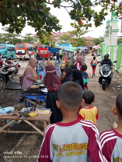 A bustling scene unfolds, featuring a vendor with a hijab preparing food near a table displaying colorful treats. Several children in school uniforms are visible, some with backpacks, heading in different directions. Behind them, a scooter rider passes by, and a row of parked buses adds to the lively atmosphere. Street stalls line the path leading towards a green and white structure, possibly a mosque. The photo is taken under trees, with camera timestamp and model information visible in the lower-left corner.
