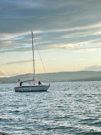 A sailboat is on the water with a mountainous backdrop and cloudy sky. There are three people on the sailboat, two visible on the deck and one sitting near the bow. The sun is setting, casting a warm glow on the clouds and water. The water is calm with small ripples. The sailboat appears to be a racing yacht, with a tall mast and sleek hull. The words "OYASEA" are visible on the stern of the boat, and "AM1371RUB578" is on the side. The image was taken in Turgoyak, Russia.