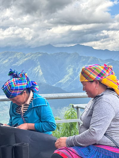 Two adult females are seated outdoors on an elevated platform with a panoramic view of mountains, likely in Tu Le, Vietnam. A grey metal railing spans horizontally between the subjects and the background.
The woman on the left wears a vibrant, multi-colored plaid headscarf predominantly featuring blue, green, and red hues, tied high with decorative tassels. She has a gold drop earring in her visible ear and a gold-colored bracelet on her right wrist. She is dressed in a light-colored striped shirt beneath a teal long-sleeved jacket. Her head is bowed as she focuses on a piece of black fabric or material laid out on a surface in front of her, holding a white tool, possibly scissors, in her right hand. Two rolls of black material are visible to her left on the same surface.
The woman on the right wears a bright, multi-colored plaid headscarf with yellow, orange, and purple as dominant colors, featuring fringes at the back. She has a silver hoop earring in her visible ear and is wearing a grey long-sleeved top with a subtle patterned texture on the shoulders. Below her waist, a traditional patterned skirt in shades of pink, blue, and red is visible. She is looking to her left with a slight smile on her face.
In the background, a vast expanse of lush, green mountains recedes into a hazy blue distance under a partly cloudy sky. Patches of white and grey clouds are interspersed with visible blue sky. Green foliage is present behind the railing between the two women.