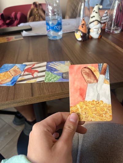 In Donji Pobori, Montenegro, a person's hand is holding a card with a watercolor painting of bread and stacks of coins. The card is in the foreground, with a table and other cards visible in the background. The table has a wooden surface, and on it are several other cards with watercolor illustrations: one depicts a box filled with golden food, another shows a red antelope against striped background, and a third illustrates a hand reaching for money. A bottle of "Rosa" water is also on the table, along with decorative gnomes. The environment suggests an indoor setting, possibly a cafe or restaurant, with a blurred background showing seating and other patrons.