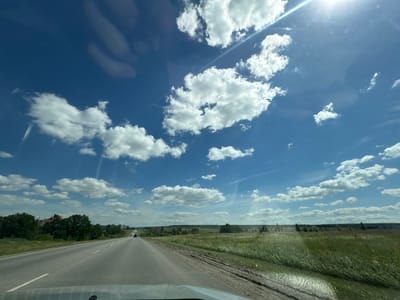 A wide-angle, slightly elevated view captures a bright, sunny day from inside a vehicle, looking forward along a paved road that curves gently to the left. The road is flanked by grassy verges and fields of green grass and some vegetation. The sky is a vibrant blue, filled with numerous fluffy white cumulus clouds. The sun is high in the sky, casting a bright glare and some lens flare. In the distance, along the left side of the road, there are trees and buildings visible, suggesting a rural or suburban setting. A few vehicles are seen on the road ahead. The overall impression is one of travel on a clear, summer day.