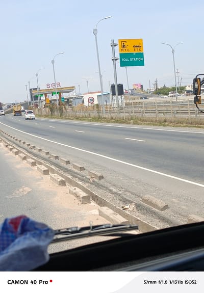 A road shot from inside a vehicle shows a multi-lane highway with a toll station sign in the distance. The sign reads "MTC ETC TOLL STATION" in yellow letters on a green background. The SGR toll station is further away. Various vehicles are on the road. There's a concrete barrier on the left side of the road. The vehicle's windshield and a partial view of a cloth are visible at the bottom of the frame. The camera information, "CAMON 40 Pro," is displayed, as is the focal length, aperture, shutter speed, and ISO: "57mm f/1.8 1/1311s ISO52."
