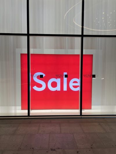 This is a nighttime exterior shot of a storefront window display. The window is divided into four panes by a black frame. Inside the window, a large red sign with white lettering announces "SALE". Below the word "SALE" are smaller white text boxes that read "SHOP ONLINE" followed by a QR code and "TH.HM.COM". The background of the display features vertical white lines. The exterior sidewalk is made of large, flat stone tiles. There are no people visible in the image.