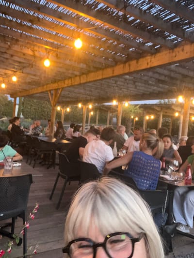 The image depicts an outdoor dining area in Andrézieux-Bouthéon, France, likely at a restaurant. Several people are seated at tables under a wooden pergola structure adorned with string lights. Diners are engaged in conversation and appear to be enjoying a meal together. The foreground features a person with blonde hair and glasses, possibly taking the photograph. Tables are set with glassware and bottles, indicating a formal dining setting. The overall atmosphere suggests a relaxed and convivial social gathering. The background reveals green foliage, suggesting an outdoor or garden setting.