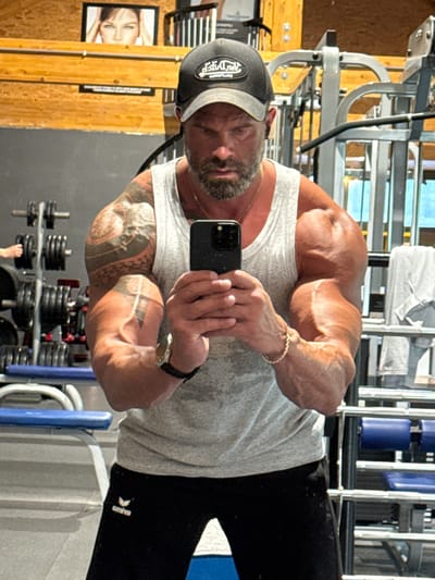 A muscular man is captured in a mirror selfie within a gym setting. He is facing the mirror, holding a black smartphone (likely an iPhone) with both hands to take the picture. He appears middle-aged with a neatly trimmed beard and is wearing a dark baseball cap, a light grey tank top, and black athletic pants. The logo "GYM LOGIC" is visible on his cap, and a white "erima" logo with its characteristic swoosh is on his pants. His highly developed arms display prominent veins and extensive tattoos. A gold chain is visible around his neck, and a gold bracelet adorns his right wrist, while a watch is on his left wrist.
The background reveals typical gym equipment, including metal weight racks with various weight plates, and exercise machines with blue padded sections. The gym's walls feature both natural wood paneling and large mirrored surfaces. On the wooden wall, a framed picture of a woman's face is visible. The lighting is bright and even, consistent with an indoor workout environment. The man's expression is focused and neutral as he takes the photograph. The overall scene suggests a dedicated fitness enthusiast. Although not visually confirmed in the image, the context indicates the location is Jemelle, Belgium.