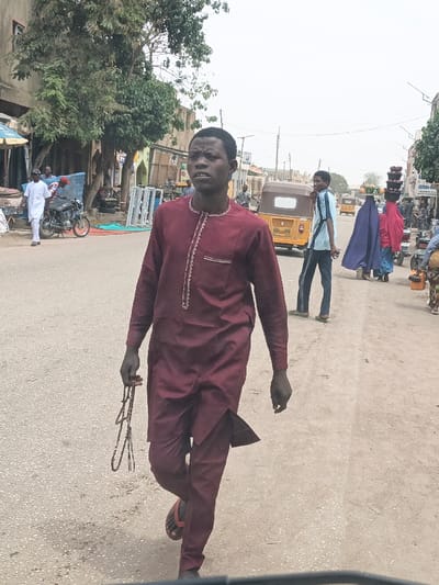 A street-level image captured in Hotoro North, Nigeria, depicts a bustling urban environment under an overcast sky. The foreground is dominated by a dark-skinned male, approximately 20-30 years old, walking towards the right side of the frame. He wears a maroon long-sleeved tunic with white embroidery around the collar and chest pocket, matching trousers, and black flip-flops. In his right hand, he holds a string of brown prayer beads. His mouth is slightly open, and his gaze is directed forward.
In the midground, several individuals and vehicles are visible. To the left, a male in a white traditional garment walks past a parked dark motorcycle. Behind them, storefronts display merchandise, partially obscured by a tree with green foliage. Centrally, a yellow and brown three-wheeled auto-rickshaw (keke Napep) is parked. To its right, another male in a light blue striped shirt and dark trousers stands facing left. Further right, two females, both with items balanced on their heads (possibly produce in baskets), walk away from the camera. One wears a dark blue full-body covering over a light blue garment, and the other a red full-body covering over a pink garment. A third person, partially visible on the far right, also carries items on their head.
The background features additional buildings lining both sides of the dusty road, some with faded paint or concrete finishes. Overhead, utility poles and electrical wires are discernible against the pale sky. The ground surface is rough and unpaved in sections, indicating a well-trafficked thoroughfare.