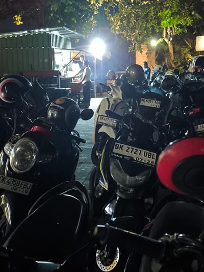 At night in Uluwatu, Indonesia, a dense parking area is packed with motorbikes under dim artificial light and leafy trees. A food stall operates in the background, where two figures appear to be cooking or preparing food. Several motorbikes have visible license plates. A parked car is visible in the distance. The area has a bustling, active atmosphere, suggesting a popular gathering spot.