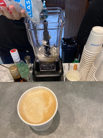 A close-up shot shows ice being poured from a bag labeled "CLASSIC" into a blender filled with liquid and ice. A hand holding the bag of ice is visible at the top left of the frame. Beside the blender are two bottles, one with a white label that reads "BACARDI" and "WHITE RUM," and another with a dark liquid and a gold cap. A small metal jigger is also present. In the foreground, a white paper cup with a frothy, tan beverage sits on a gray, textured surface. To the right of the blender, a stack of white paper cups with blue text that appears to read "TOPSHOP" is visible, along with a single white cup containing clear liquid. A black trash bag is tucked behind the blender. The background features wooden paneling and a hint of a person wearing black clothing. The scene appears to be a bar or kitchen setting where a drink is being prepared.