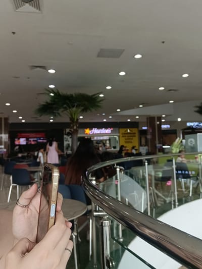 The image depicts a brightly lit indoor space, likely a food court or shopping mall atrium, as indicated by the presence of various food establishments and ceiling lights. A woman's hands are visible in the foreground, holding a gold-colored smartphone. In the background, several people are seated at tables and walking around. One woman in a pink dress stands near a palm tree. Signs for "Hardee's" and other businesses are visible on the storefronts. The environment appears to be a modern commercial setting.
