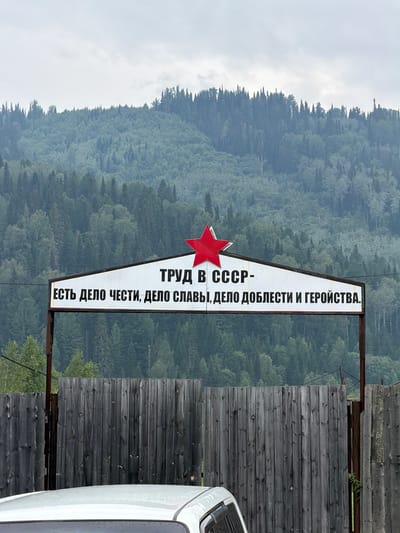 An outdoor, eye-level, medium shot captures a sign with a red star above a white background with black Russian text. The text reads "ТРУД В СССР - ЕСТЬ ДЕЛО ЧЕСТИ, ДЕЛО СЛАВЫ, ДЕЛО ДОБЛЕСТИ И ГЕРОЙСТВА." translated to "LABOR IN THE USSR IS A MATTER OF HONOR, A MATTER OF GLORY, A MATTER OF VALOR AND HEROISM." The sign is mounted on poles and appears to be a historical monument or propaganda sign. Behind the sign, a dense, misty forest covers a mountain slope under a cloudy sky. In the foreground, a weathered wooden fence runs horizontally across the image, partially obscuring the lower portion of the sign. The roof of a white car is visible in the bottom left corner. The image was taken in Ust'-Kabyrza, Russia.