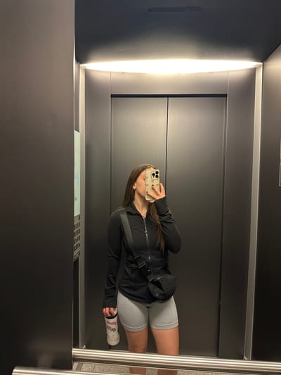 A woman is taking a selfie in an elevator. She is wearing a black zip-up jacket, grey shorts, and a white bandage on her left wrist. She is holding a phone with a floral-patterned case. The elevator has grey walls and a metal control panel. It is illuminated by a bright light panel. It is not possible to verify the location.
