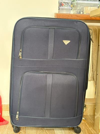 The image shows a dark blue suitcase with multiple zippers and a triangular logo on the front. The suitcase is standing upright on a wooden floor, with a light-colored wall behind it. To the right of the suitcase, there is a beige towel hanging and part of a closet. On a shelf above the suitcase, several clear plastic storage containers are visible. The scene appears to be indoors, possibly in a home or storage area. The specific location in Baku City, Azerbaijan, is not determinable from the image.