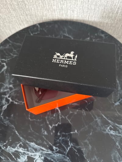 A black Hermès box is open, revealing a pair of pink sunglasses inside. The box is placed on a dark marble surface. The background is a textured, light-colored fabric. The image was taken in the city of Soloneshnoye, Russia.
