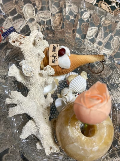 In the city of Margorejo, Indonesia, a close-up, overhead shot showcases a decorative arrangement within a clear, faceted glass bowl. The focal point is a collection of beach-themed items: a piece of white coral, several small seashells, a larger shell, and a ring. A small, dark red bead is affixed to a cluster of white shells. Attached to the larger shell is a small, wooden tag with the handwritten words "Thank You". Partially visible in the bottom right is a donut-shaped, yellowish object topped with a pale orange, rose-shaped ornament. The background of the bowl reveals reflections of a patterned fabric or surface.