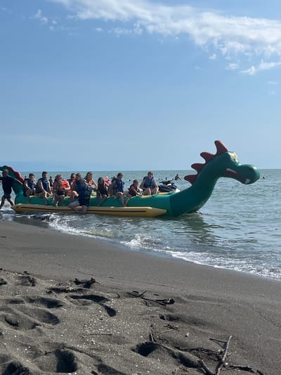 This image shows a group of people on an inflatable dragon-shaped banana boat at the edge of the Black Sea in Ureki, Georgia. The boat is green with a red snout and has a long, segmented body. Several people, wearing life vests in various colors like blue and orange, are seated along the length of the boat. They appear to be enjoying a sunny day at the beach, with the water lapping at the shore.

The scene is set on a dark, sandy beach with small shells and pieces of driftwood scattered on the sand. The water is clear and calm, with gentle waves washing ashore. The sky is bright blue with a few fluffy white clouds, indicating good weather and likely daytime. The overall mood seems relaxed and fun, with people preparing for or just finishing a water activity. The inflatable dragon's head is positioned at the front of the boat, emerging from the water.