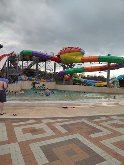 The image depicts a water park in Shcherbani, Ukraine, under an overcast sky. In the foreground, a patterned brick walkway transitions to a sandy area with several pairs of flip-flops scattered on it. Beyond the sand, a shallow pool with wavy water is visible, populated by several people swimming and wading. A man wearing blue shorts stands in the pool, facing away from the camera. Behind the pool, a large, multi-colored water slide structure dominates the scene. The slide system is comprised of various colored tubes, including red, purple, green, and orange, converging and diverging in a complex arrangement. Some parts of the slide are supported by metal scaffolding. To the left of the main slide structure, a red water slide with white accents is also visible, leading down from a platform. Several inflatable rings are placed near the edge of the pool. In the background, trees and foliage are visible, suggesting a natural landscape surrounding the park. The overall atmosphere appears to be one of leisure and recreation.
