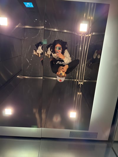 A couple takes a selfie in a modern elevator with reflective walls and ceiling. The woman has long dark hair and is wearing a light-colored sweatshirt, while the man has a baseball cap and a dark sweatshirt. The elevator has a sleek, dark marble floor and minimalist lighting fixtures. A control panel is visible on the left, and ventilation grates are on the right. Their reflections appear on the ceiling and floor of the elevator.