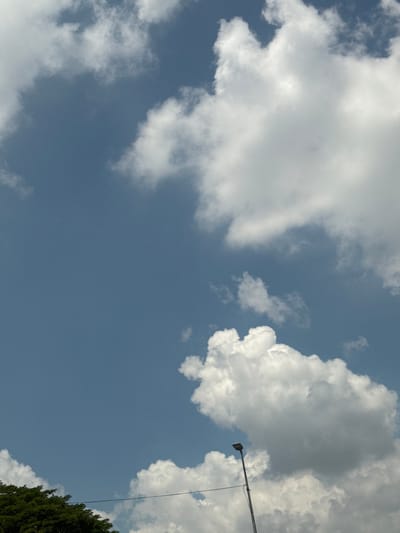 The image shows a clear blue sky with scattered white cumulus clouds. The perspective is looking upwards, with a portion of a tall, green tree visible on the right side of the frame, suggesting an outdoor setting. A street lamp mounted on a pole is also visible on the right, with a wire extending upwards from it. The lighting indicates it is daytime, with the sun high in the sky. The presence of cumulus clouds suggests fair weather, possibly with a gentle breeze. The location context provided is Kuala Lumpur, Malaysia. There is no visible text in the image.