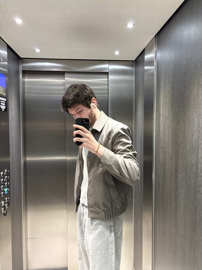 An adult male is observed inside an elevator located in Cachan, France. The individual, possessing dark brown hair and a short beard, is depicted from mid-thighs upwards. He wears a light-toned collared jacket over a light grey shirt and light-colored trousers. A white wireless earbud is present in his right ear. On his left wrist, a gold-colored cuban link chain bracelet is visible, and a gold-colored ring adorns his left ring finger. He holds a black smartphone in his left hand, positioned to capture his reflection, indicating a self-portrait action. His gaze is directed downwards towards the smartphone. The elevator interior features brushed stainless steel panels on the left wall and the rear door, while the right wall is covered with a dark, textured panel. The ceiling is white and fitted with two recessed circular lights. An elevator control panel, displaying multiple circular buttons and a digital screen, is affixed to the left wall.
