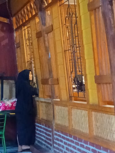 The image captures a side view of a building in Ciamis, Indonesia, where a person is standing. The building's exterior features a blend of wooden architectural elements, including vertical panels in yellowish and light brown hues, and sections with a lighter, possibly woven or textured, pattern below the windows. The base of the wall is constructed with a red brick pattern. Several windows are visible, each adorned with ornate, dark metal grilles. One window on the right provides a glimpse into the interior or reflects a dark object.

The main subject is a person, likely a woman, positioned on the left side of the frame. She is dressed in a long, black garment and a black hijab, covering her head and neck. Her face is seen in profile, with a serene expression, as she looks towards the right, possibly observing something through the window or beyond the frame. Her right arm is slightly raised, suggesting an interaction with the window area. Her bare feet are visible beneath her attire.

To the far left of the person, a portion of a green, possibly metal, stool or small table is visible. On this surface rests a white and pink object, which appears to be a plastic bag or a box of tissues. The lighting in the image suggests it is daytime, potentially with indirect light. The architectural style and the person's traditional attire are consistent with the provided location context of Ciamis, Indonesia. There is no visible text present in the image.