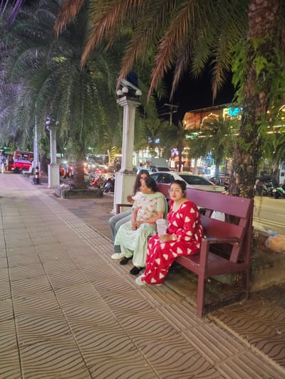 Three people are sitting on a bench at night, with palm trees and a street scene in the background. The person on the left is wearing a red and white patterned pajama set. She is wearing glasses and holding a clear plastic cup. The person in the middle is wearing a light green and white floral print top and light gray pants. The person on the right is wearing a light green patterned top and gray pants. The bench is made of dark red wood. The ground is paved with textured tiles. The background shows a street with buildings, lights, parked motorcycles, and a red vehicle that appears to be a truck or tuk-tuk. The scene suggests a tropical location at night, possibly a street market or tourist area. The lighting indicates it is nighttime, and the presence of palm trees points to a warm climate. There is no visible text in the image.
