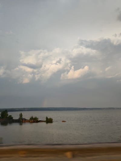 The image shows a body of water, possibly a lake or river, under an overcast sky with clouds. In the distance, there is a land mass with a visible horizon line. A small, tree-covered island is located in the water. The location is Пальники, Russia. A faint rainbow is discernible in the sky. The foreground is out of focus, suggesting the image was taken from a moving vehicle or behind an object. No people are clearly visible.