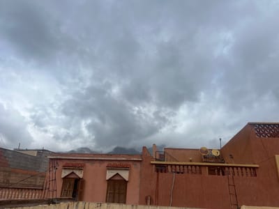 The image depicts a section of Ouaouizert, Morocco, under an overcast sky. The foreground is dominated by the rooftops of buildings constructed with a reddish-brown textured stucco. On the left, a building made of gray cinder blocks is visible. Adjacent to it stands a two-story building with wooden shutters and a tiled roof section above each window. To the right, a building with two satellite dishes is on its roof behind a balcony. A ladder is leaning against the wall of this building. In the background, mountains are shrouded in mist.