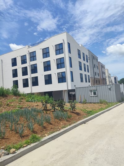 The image depicts a newly constructed, modern white building with multiple floors and dark-framed windows, located in the city of Asan-si, South Korea. The building is situated on a slight incline, with a patch of cultivated greenery, including rows of scallions, in the foreground. A grey shipping container-style structure with a small window is positioned alongside the building. The sky is clear blue with scattered white clouds, suggesting a daytime scene. The surrounding area appears to be under development, with a gravelly path and a concrete curb visible. There are no people or visible signs of activity in the image.