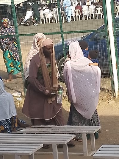 This image depicts an outdoor gathering in Sabon Gari East, Nigeria. In the foreground, a light-colored slatted bench is partially visible, with a person seated next to it, wearing a light headscarf and patterned skirt.

The central focus includes three women standing in the midground. One woman, with a darker complexion, wears a light brown hijab and a long-sleeved brown tunic, holding a clear plastic water bottle in her right hand and a small, dark object in her left. She is partially obscured by another woman wearing a patterned headscarf directly behind her. To their right, a third woman faces away from the viewer, clad in a light, patterned head covering and a dark, patterned skirt. She holds two small children, both wearing dark beanies and blue clothing.

A green wire mesh fence separates the midground subjects from the background. Behind this fence, a dark-colored sedan is parked. Further in the background, numerous individuals, primarily men in light-colored traditional attire and head coverings, are seated on white plastic chairs. A person in light blue stands among them, and a bicycle is visible near the seated group. The ground appears dry and dusty under daylight conditions.