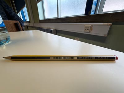 A single yellow pencil with black stripes, sharpened at its left end and featuring a red-capped right end, rests horizontally on a smooth, light-colored tabletop. Visible markings on the pencil include a barcode, "EAN 40 07817 104620," and "Art. Nr. 120-2." To the upper left, a clear plastic water bottle with a blue label and cap is partially visible. The background reveals a window structure with multiple panes, through which ambient light enters. Below the window, a white horizontal panel shows signs of wear and chipping, exposing a darker material underneath. A dark green wall is visible beneath this panel. A wooden chair back, draped with dark fabric, is partially discernible on the far left. The scene depicts a static interior room, consistent with a classroom or study area, located in Vila Real, Portugal. No individuals are present in the image.