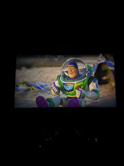 This media file shows a close-up, vertically oriented shot of a Buzz Lightyear action figure. The toy is in a crouching position, with its left arm extended and its right hand near its chest. Buzz is wearing his iconic white and green spacesuit with purple accents on his gloves and boots. His helmet is clear, revealing his cartoonish face with wide, surprised eyes and a slightly open mouth. The word "LIGHTYEAR" is printed in white text on a green label on his chest. The background is blurry and appears to be a textured, ground-like surface, possibly sand or dirt, with some blue and yellow elements suggesting debris or other toys. The lighting is dramatic, with highlights on Buzz's suit and a spotlight effect illuminating the figure. The overall mood is one of action or surprise. The location context is Wichit, Thailand, but this is not directly visible in the image.