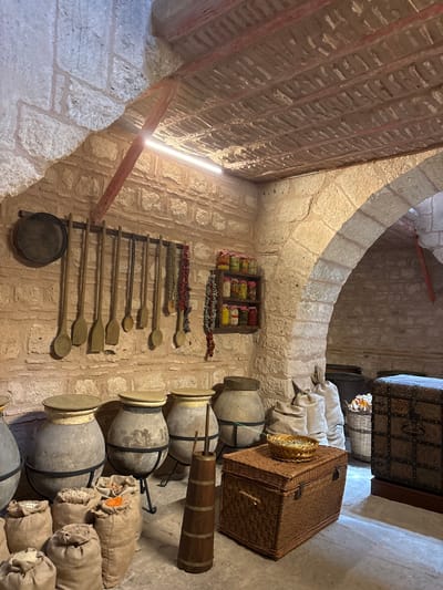 The image depicts an interior scene of what appears to be a traditional Turkish storage area or shop in Istanbul, Turkey. The setting is characterized by rough-hewn stone walls and arched architectural features, suggesting an old building. Various objects are displayed, hinting at a historical or artisanal context.
Prominently featured are several large, grey ceramic jars, some with lids, stacked in a row. Next to them are numerous sacks filled with what appear to be grains or dried goods, with some sacks showing hints of orange and white contents. On a wall to the left, a series of wooden spoons and spatulas of varying sizes are neatly arranged on a rack. Above the spoons, a small wooden shelf holds jars filled with colorful dried goods, possibly spices or preserved fruits, and a strand of dried chili peppers hangs below.
In the background, a large, ornate wooden chest with metal accents rests on a surface. Next to it is a woven basket, and a large, woven picnic-style basket is also visible in the foreground. A metal sieve or colander is placed on the wall near the bottom left. The lighting suggests it could be daytime, with a bright light source casting shadows and highlighting the textures of the stone and objects. The overall impression is one of a well-preserved historical space showcasing traditional storage methods and goods. There is no visible text in the image.