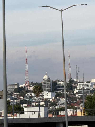 The image features an urban landscape with multiple buildings and towers. A tall, red-and-white striped communications tower is prominent. Other towers of varying heights are visible in the distance. Residential and commercial buildings are densely packed together. A curved, gray lamppost is in the foreground on the left. The sky is overcast.