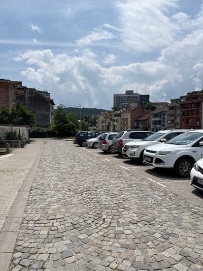 The image depicts an urban street scene in Gabrovo, Bulgaria, during daytime under a partially cloudy sky. The foreground is composed of a cobblestone street featuring irregular grey paving stones arranged in a fanned pattern, bordered by a lighter grey paved sidewalk on the left. In the midground, a series of vehicles is parked diagonally along the right side of the cobblestone street within delineated white parking lines. From left to right, these include a dark-colored minivan/SUV, a silver Nissan Qashqai-like SUV, a white Nissan Qashqai-like SUV with a visible Bulgarian license plate "EB 7244 BH", a white Ford Kuga-like SUV with a partially visible Bulgarian license plate, and the front portion of another white vehicle with a Bulgarian license plate beginning "EB".

Behind the parked cars, a diverse architectural landscape is visible. On the left, older, multi-story buildings with brick and grey facades appear partially dilapidated and are partially obscured by dense green trees and vegetation. To the right, a row of more brightly colored multi-story residential buildings with various architectural details and visible air conditioning units line the street. In the distant background, a tall, modern building with a grey and dark facade stands against the sky. Further right, green, tree-covered hills are visible beneath a darker cloud formation. The sky is largely bright with scattered cumulus clouds. No individuals are present within the frame.