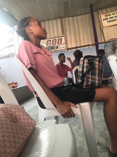 A woman wearing a pink collared shirt and dark bottoms sits on a white plastic chair. A plaid bag is next to her. In the background are other people seated. Religious signs are affixed to the wall including "WHO GOD SPECIMEN" and "SEEK YE E AND HIS RI THINGS". The room appears to be a church or similar gathering space, with corrugated metal roofing visible. A pink purse is visible on the right side of the photo.