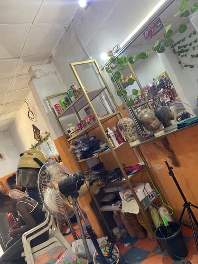 This image depicts the interior of a beauty salon, possibly located in Du, Nigeria, given the context. The scene is brightly lit by overhead lights. On the left, a woman sits in a white plastic chair, her head bowed as if receiving a hair service. Beside her, a large, cream-colored hair dryer hood is positioned over where a client's head would be. In front of the woman and to her right, a silver oscillating fan is on.

Against the back wall, a gold-colored metal shelf unit displays various hair products, wigs, and mannequins. The shelves hold an assortment of haircare products in various containers, including bottles and jars. Several wigs are neatly arranged on the shelves, some in plastic packaging. Two mannequin heads, one with a floral pattern and the other with a smooth finish, are placed on a shelf, with the face of one clearly visible. Further back, more wigs and hair products are visible, suggesting a well-stocked salon.

The walls are painted in shades of orange and white, with some visible signs of wear. The floor has a tiled pattern in blue and orange. A black tripod with a light fixture is set up on the right side of the image. There are also several plastic buckets and bottles scattered around the floor, along with a basket that appears to contain hair rollers. A sign with green leaves and a picture of a woman is visible hanging from the ceiling, possibly advertising the salon. The overall impression is of a busy and functional hair salon.