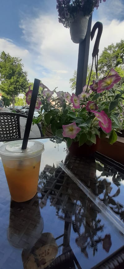 A clear plastic cup filled with an orange liquid and a black straw sits on a reflective glass table. In the background, pink and yellow petunias bloom in a planter, with a blurred street scene and trees visible beyond. The location is Yessentuki, Russia, a resort city known for its mineral springs and spa culture. The scene suggests a cafe or outdoor seating area, possibly a place for tourists to relax and enjoy a beverage while taking in the surroundings. The reflection on the table shows the intricate pattern of the table itself, as well as the flora and sky above, adding depth and visual interest to the composition.
