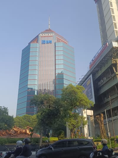 In Surabaya, Indonesia, a tall, modern skyscraper with a glass facade dominates the frame, its blue and orange panels reflecting the clear sky. The building is topped with a spire and features the "PLAZA BRI" and "BRI" logos in blue letters. To the right of the skyscraper, another building rises, partly obscured by scaffolding and a large billboard advertising a car. In the foreground, lush green trees line a street where several motorcycles and a car are visible. On one of the motorcycles, two individuals wearing helmets are riding. Further left, a bronze-colored statue depicting a scene of struggle or movement is situated amidst greenery. The overall scene depicts an urban environment with contemporary architecture, transportation, and public art.