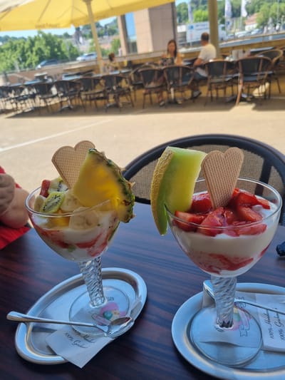 The image shows two fruit sundaes on a dark wooden table, outdoors. One sundae contains strawberries and melon, and the other contains pineapple, kiwi, and banana. Each sundae has a heart-shaped wafer. The sundaes are served on silver trays with spoons and napkins. In the background, there are tables and chairs of an outdoor café, partially under a yellow sunshade. Across the street, there are buildings, trees, and people. The scene appears to be set in an urban area, potentially the city of Saarbrücken, Germany.