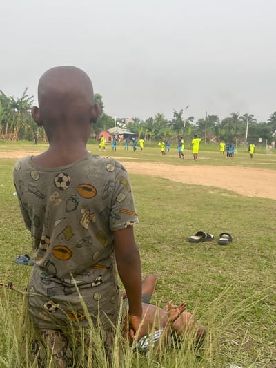 The image depicts a young boy sitting on the grass in a field in Ikot Ekpene, Nigeria. He is wearing a t-shirt with various sports-related graphics. Tall grass obscures part of his body. In the background, a group of people dressed in athletic attire appear to be playing or practicing a sport on a dirt field, possibly soccer or a similar game. There are trees and other vegetation visible in the background. A pair of sandals are lying on the grass between the boy and the playing field. The sky is overcast.