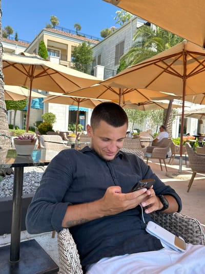 A young man is seated outdoors at a cafe in Donja Lastva, Montenegro, looking down at his smartphone and holding it with both hands. He is wearing a dark blue collared shirt. A white portable charger is resting on his lap, connected to his phone. He is sitting in a wicker chair, with a table to his left. In the background, there are several large beige umbrellas providing shade over other tables and chairs. The architecture suggests a resort or hotel setting, with buildings featuring light-colored facades and terracotta roofs. Palm trees and lush greenery are visible, indicating a warm climate. The sky is clear and blue.
