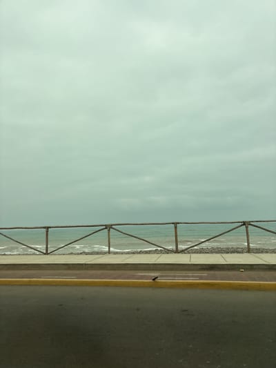 The image is a photograph of a coastal scene in San Miguel, Peru. The main subjects are the ocean, a pedestrian walkway, and a road. The ocean is on the left side of the image, with gentle waves lapping against a rocky shore. A metal railing, possibly a barrier, runs along the edge of the walkway, separating it from the sea. The walkway is paved with light-colored material and has a darker, reddish-brown strip running through it, with faint white lines, possibly indicating lanes or a path. Beyond the walkway is a road, paved with asphalt and bordered by a yellow line. The sky is overcast and a pale, muted blue-green color, suggesting a cloudy or hazy day. There is no apparent activity or event taking place; the scene appears to be a quiet, still moment. The time of day is difficult to determine precisely due to the overcast conditions, but it does not appear to be dawn or dusk. The overall mood of the image is serene and somewhat melancholic, with the subdued colors and lack of human presence. No text is visible in the image.