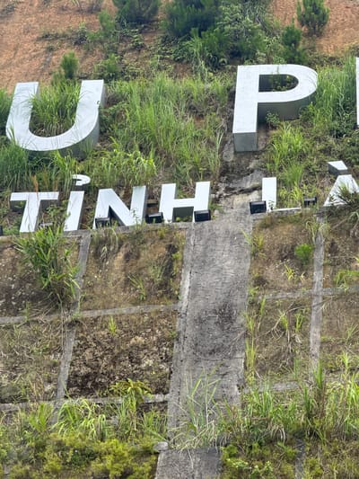 The image displays a portion of a hillside in Tu Le, Vietnam, featuring large, three-dimensional block letters embedded into the terrain. The upper visible line of text clearly shows the letters "U" and "P" in a light silver or grey finish. The lower visible line of text reads "TINH LA", rendered in the same style of letters, with two black, rectangular spotlights positioned in front of the letters "H" and "A".
The hillside itself is characterized by significant natural overgrowth. Lush green grass and various small plants densely surround the letters, with some vegetation growing within the hollows of letters like the "U" and "P". Patches of reddish-brown earth are visible between the vegetation, particularly higher on the slope. Below the letters, the terrain is structured with grey concrete retaining walls or steps, which form a grid-like pattern in the lower sections of the image. These concrete structures are also partially covered with moss, dirt, and green foliage, indicating exposure to the elements and natural integration into the environment. No people or animals are visible in the scene.