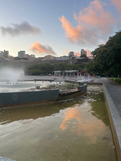 The image depicts an outdoor public space in Las Palmas, Spain, featuring a prominent fountain in the foreground with several water jets active. The fountain is surrounded by a shallow body of water with a concrete edge. In the middle ground, a playground area is visible with colorful structures, including slides and a fenced-in section. Benches are placed around the playground. Beyond the playground, a street or pathway runs alongside a line of trees. In the background, a series of buildings of varying heights and architectural styles are clustered on a hillside, with some appearing to be residential and others commercial, including a building with "BINGO" signage. The sky above displays soft, diffused clouds tinged with the colors of sunrise or sunset, reflecting in the water of the fountain. The overall scene suggests a public park or recreational area within the urban environment of Las Palmas.